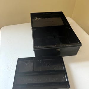 Puede incluir: Una caja registradora de metal negro abierta, con una bandeja extra&iacute;ble. La caja es rectangular con una tapa con bisagras y una peque&ntilde;a cerradura. La bandeja tiene varios compartimentos para organizar objetos. La caja est&aacute; sobre una superficie blanca.