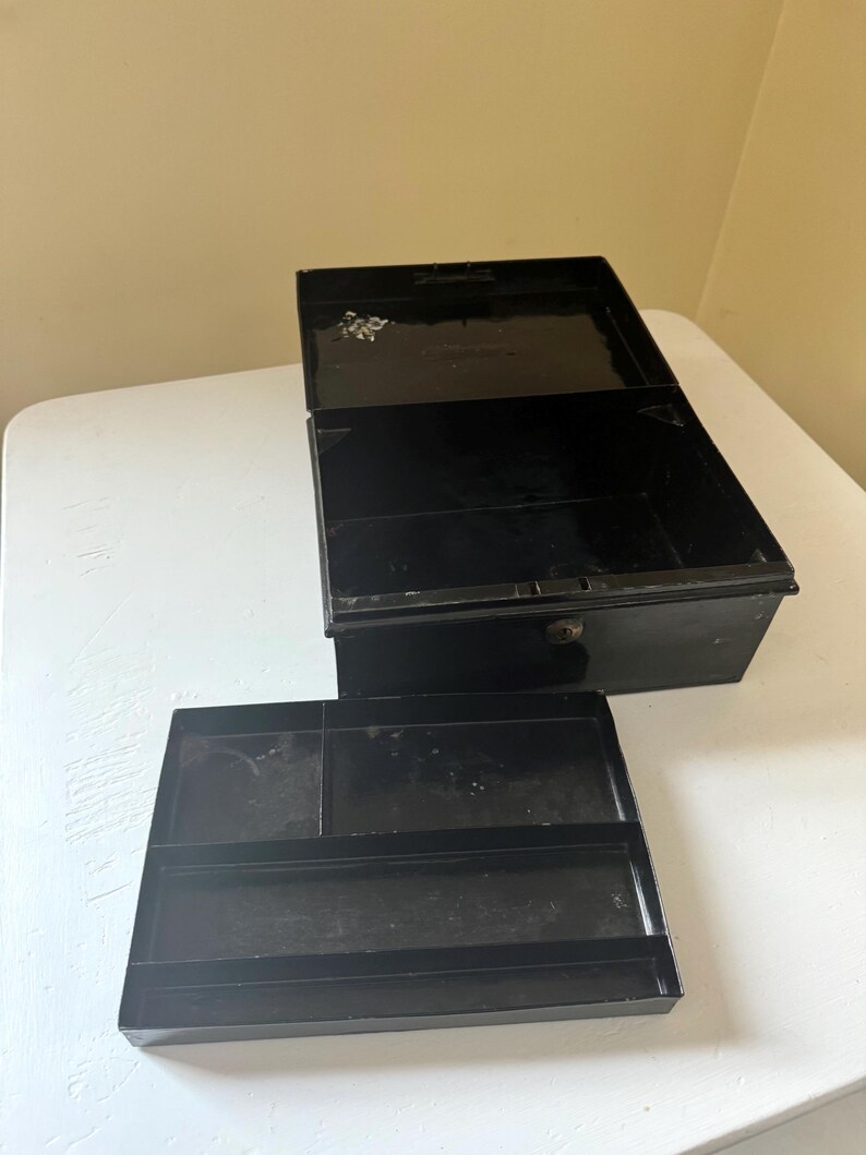 Puede incluir: Una caja registradora de metal negro abierta, con una bandeja extra&iacute;ble. La caja es rectangular con una tapa con bisagras y una peque&ntilde;a cerradura. La bandeja tiene varios compartimentos para organizar objetos. La caja est&aacute; sobre una superficie blanca.