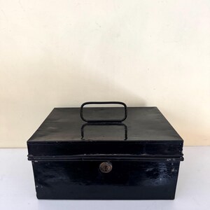 Puede incluir: Una caja registradora rectangular de metal negro con un asa en la parte superior y una cerradura en la parte delantera. La caja tiene un acabado brillante y parece ser vintage. El asa es un lazo rectangular simple.