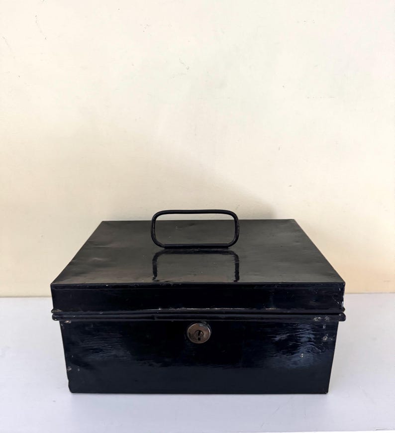 Puede incluir: Una caja registradora rectangular de metal negro con un asa en la parte superior y una cerradura en la parte delantera. La caja tiene un acabado brillante y parece ser vintage. El asa es un lazo rectangular simple.