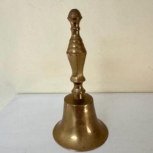 May include: A vintage brass bell with a long, ornate handle and a flared base. The bell has a polished finish, reflecting the light. The handle features a series of decorative rings and a rounded top. The bell is likely used for calling or signaling.