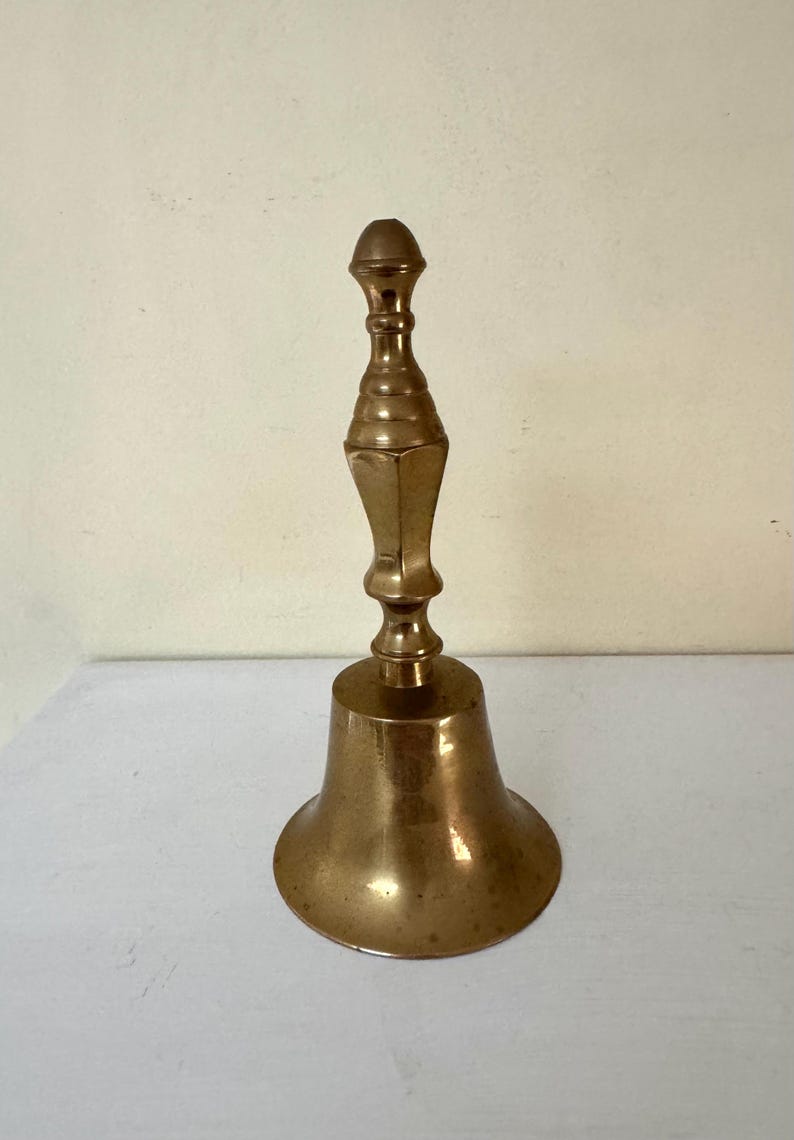 May include: A vintage brass bell with a long, ornate handle and a flared base. The bell has a polished finish, reflecting the light. The handle features a series of decorative rings and a rounded top. The bell is likely used for calling or signaling.