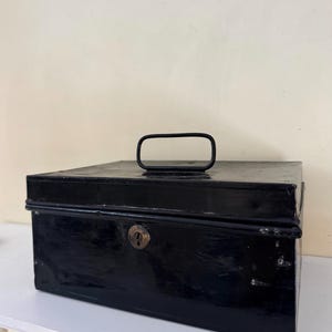 Puede incluir: Una caja met&aacute;lica negra rectangular con asa y cerradura. La caja tiene un acabado brillante y parece ser vintage. El asa es un lazo rectangular simple. La caja est&aacute; sobre una superficie blanca.