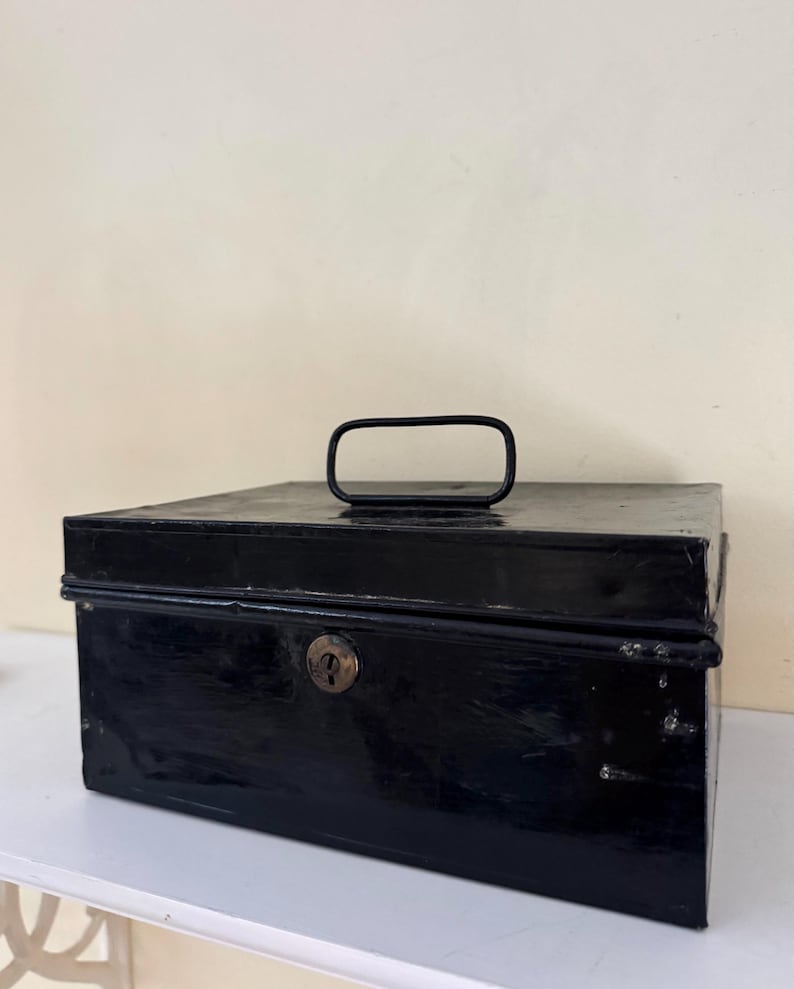 Puede incluir: Una caja met&aacute;lica negra rectangular con asa y cerradura. La caja tiene un acabado brillante y parece ser vintage. El asa es un lazo rectangular simple. La caja est&aacute; sobre una superficie blanca.