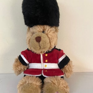 May include: A brown teddy bear wearing a red and black uniform with a black furry hat. The bear is sitting on a white surface.
