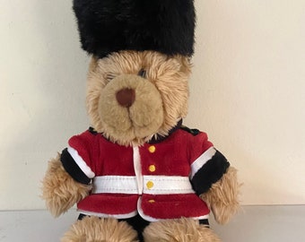 May include: A brown teddy bear wearing a red and black uniform with a black furry hat. The bear is sitting on a white surface.