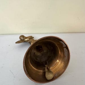 May include: A vintage brass bell with a small angel figurine on top. The bell has a rounded shape with a clapper inside. The angel is in a flying pose. The bell is likely used for decoration or as a collectible item.