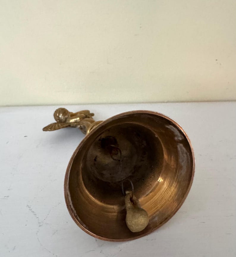 May include: A vintage brass bell with a small angel figurine on top. The bell has a rounded shape with a clapper inside. The angel is in a flying pose. The bell is likely used for decoration or as a collectible item.