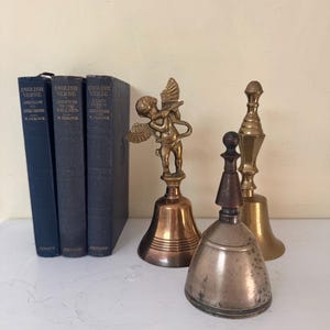 May include: An assortment of vintage bells and books. Three dark blue books with the text "ENGLISH VERSE" are on the left. Three metal bells of varying sizes and designs are on the right, including a bell with an angel figurine.
