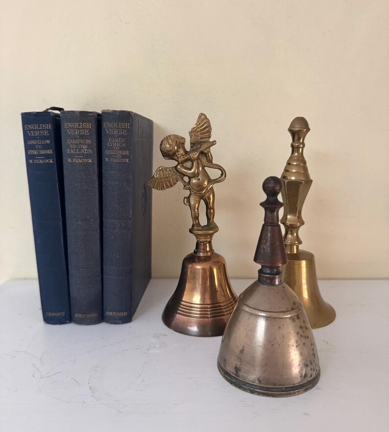 May include: An assortment of vintage bells and books. Three dark blue books with the text "ENGLISH VERSE" are on the left. Three metal bells of varying sizes and designs are on the right, including a bell with an angel figurine.
