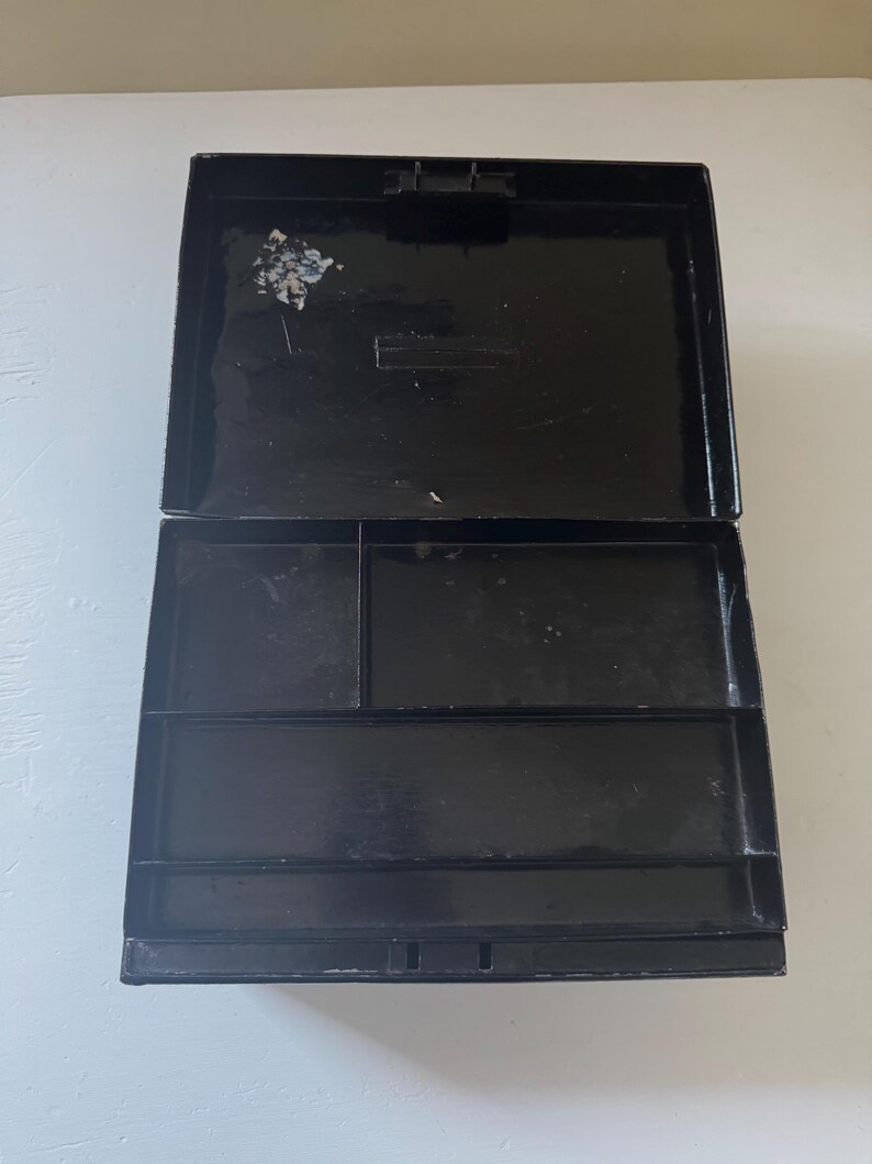 Puede incluir: Caja rectangular negra abierta, con tapa con bisagras. El interior est&aacute; dividido en varios compartimentos de diferentes tama&ntilde;os. La caja parece estar hecha de un material brillante, y la tapa est&aacute; ligeramente entreabierta.
