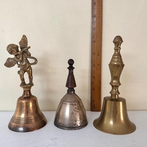 May include: Three antique bells of varying designs. The bell on the left features a cherub figure. The center bell is silver-toned with a wooden handle. The bell on the right is brass with a decorative handle. A wooden ruler is in the background.