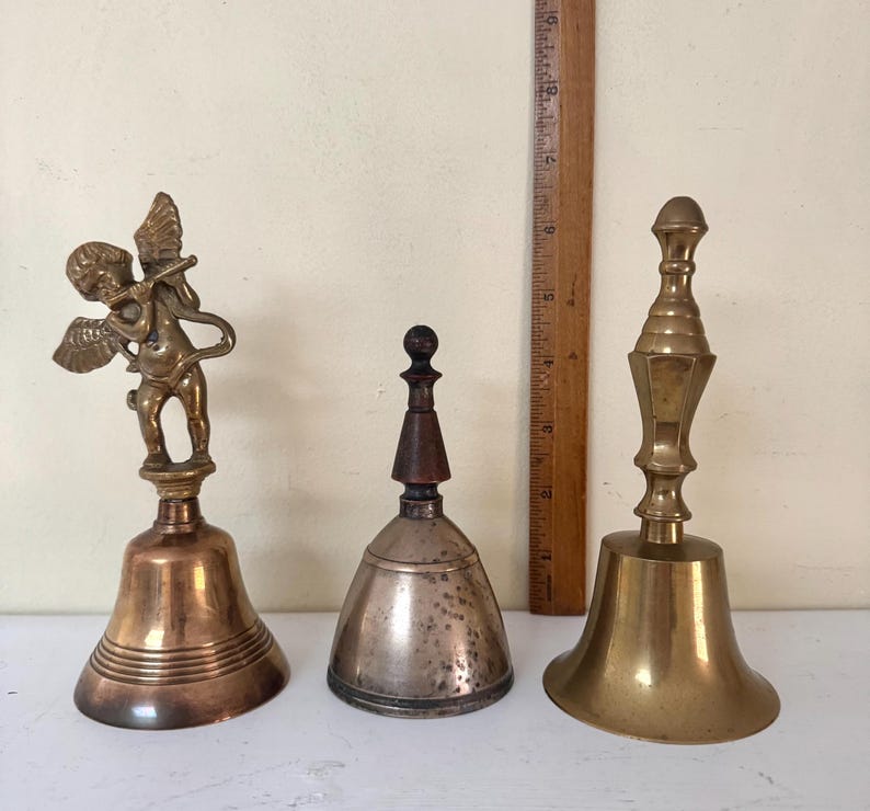 May include: Three antique bells of varying designs. The bell on the left features a cherub figure. The center bell is silver-toned with a wooden handle. The bell on the right is brass with a decorative handle. A wooden ruler is in the background.