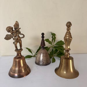 May include: Three antique bells of varying designs. The bell on the left features a small cherub figure. The center bell is silver-toned with a wooden handle. The bell on the right has a long handle and a flared base. All are made of metal.