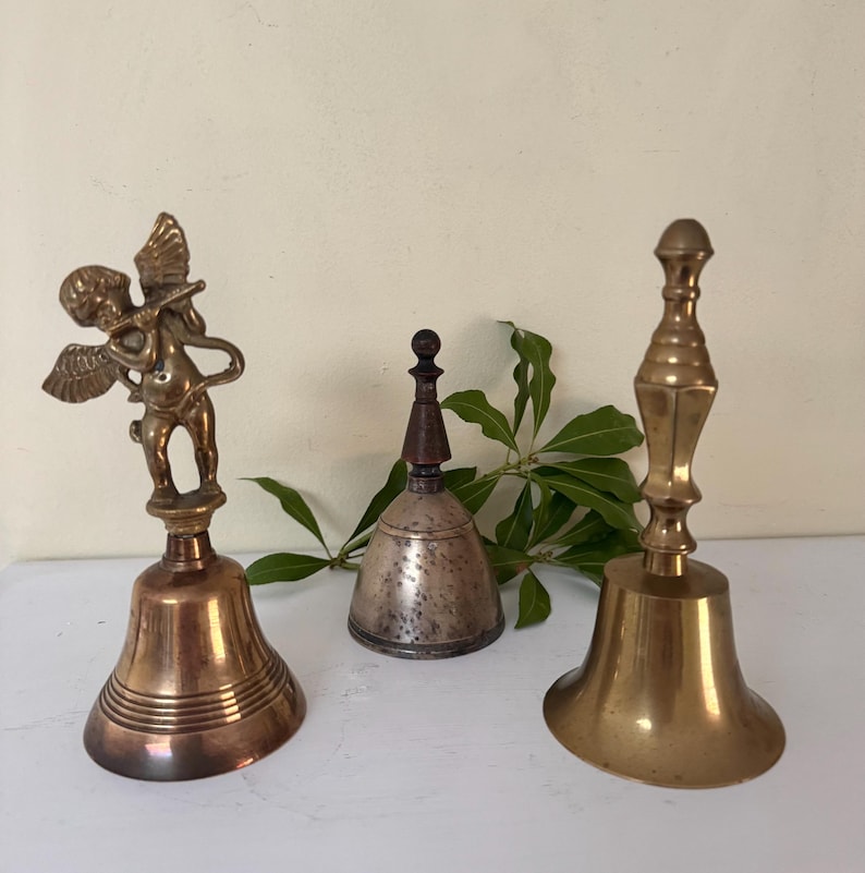 May include: Three antique bells of varying designs. The bell on the left features a small cherub figure. The center bell is silver-toned with a wooden handle. The bell on the right has a long handle and a flared base. All are made of metal.