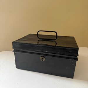 Puede incluir: Una caja registradora de metal negro con asa y cerradura. La caja rectangular est&aacute; sobre una superficie blanca. El asa es una barra curva simple.