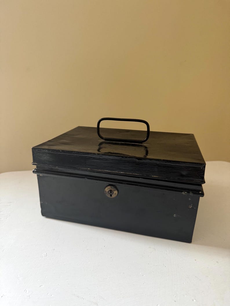 Puede incluir: Una caja registradora de metal negro con asa y cerradura. La caja rectangular est&aacute; sobre una superficie blanca. El asa es una barra curva simple.