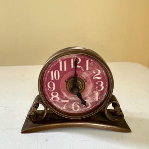 May include: A round table clock with a metallic bronze-toned frame and base. The clock face is a deep magenta color with white numerals from 1 to 12. The center of the face features the text "EDWD. MERCER LIVERPOOL" in a circular pattern.