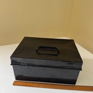 Puede incluir: Una caja met&aacute;lica negra con asa en la parte superior. La caja es rectangular y tiene un acabado brillante. Una regla de madera est&aacute; al lado de la caja. La caja probablemente se utiliza para el almacenamiento.