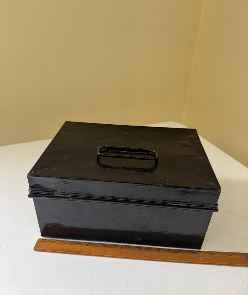 Puede incluir: Una caja met&aacute;lica negra con asa en la parte superior. La caja es rectangular y tiene un acabado brillante. Una regla de madera est&aacute; al lado de la caja. La caja probablemente se utiliza para el almacenamiento.