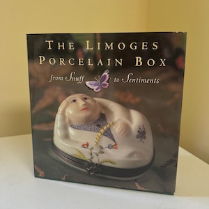 May include: A hardcover book titled The Limoges Porcelain Box from Snuff to Sentiments. The cover features a photograph of a white porcelain figurine shaped like a reclining person with painted floral details and a purple butterfly.