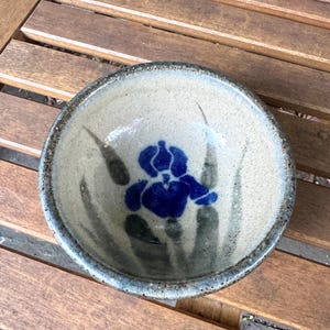 May include: A round ceramic bowl with a speckled beige glaze and a dark rim. The interior features a hand-painted deep blue iris flower centered among muted green, elongated leaf shapes that extend from the base toward the bowl's edge.