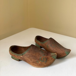 May include: A pair of wooden clogs, featuring a brown finish with green accents along the edges and heels. The clogs have a traditional design, with a rounded toe and a hollowed-out interior. The clogs are likely vintage or antique.