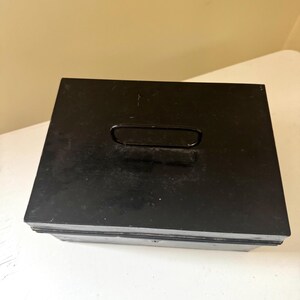 Puede incluir: Una caja de metal negro con forma rectangular y un asa en la tapa. La caja tiene un aspecto ligeramente desgastado, lo que sugiere que puede ser vintage o usada. El asa tiene un dise&ntilde;o simple y curvado.
