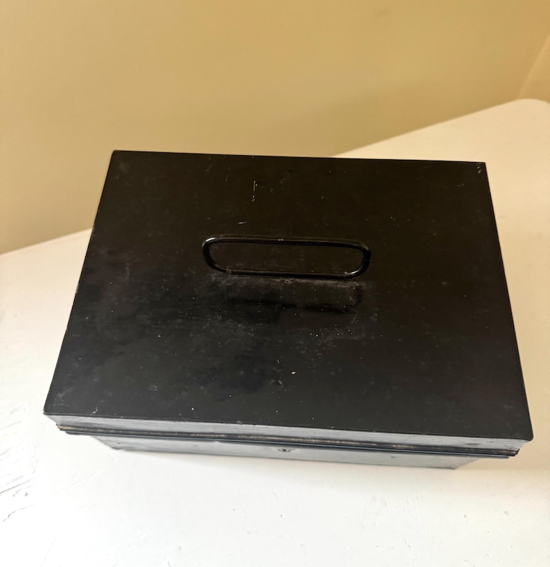Puede incluir: Una caja de metal negro con forma rectangular y un asa en la tapa. La caja tiene un aspecto ligeramente desgastado, lo que sugiere que puede ser vintage o usada. El asa tiene un dise&ntilde;o simple y curvado.