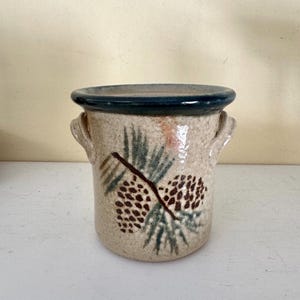 May include: A beige ceramic utensil holder with a dark blue rim and two small handles. The holder is decorated with a pine cone and pine needle design in brown and green.