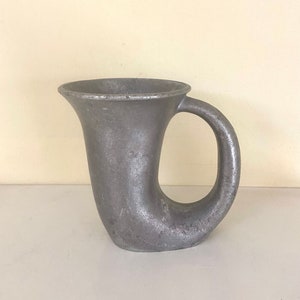 May include: A silver metal mug with a unique curved handle. The mug is made of a smooth, shiny metal and has a simple, elegant design.