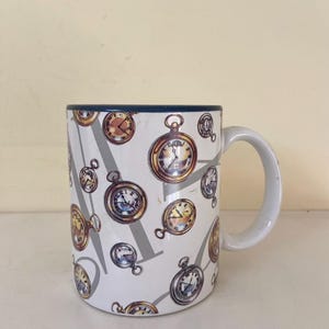 May include: White ceramic mug with a blue rim and a pattern of gold pocket watches on a gray background.