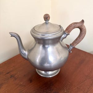 May include: A vintage pewter teapot with a wooden handle and lid knob. The teapot has a curved spout and sits on a round base. The wooden handle and knob add a touch of warmth to the otherwise cool-toned metal. A classic piece for tea lovers.
