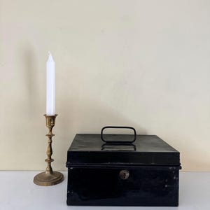 Puede incluir: Una caja registradora de metal negro con asa y cerradura de llave se encuentra junto a un candelabro de lat&oacute;n que sostiene una vela blanca. La vela no est&aacute; encendida. Los objetos est&aacute;n sobre una superficie blanca.