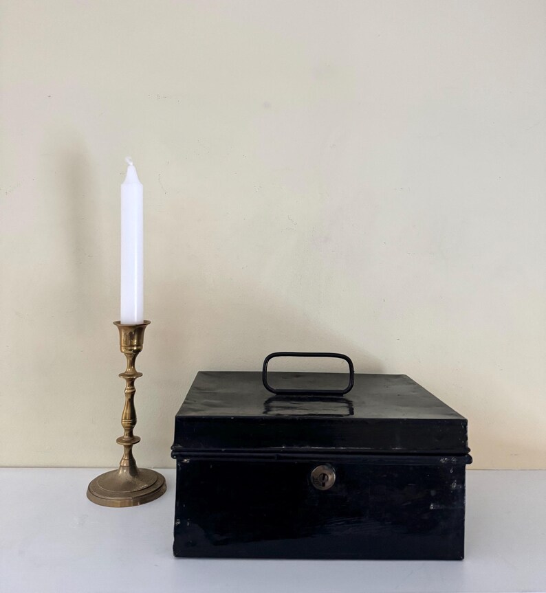 Puede incluir: Una caja registradora de metal negro con asa y cerradura de llave se encuentra junto a un candelabro de lat&oacute;n que sostiene una vela blanca. La vela no est&aacute; encendida. Los objetos est&aacute;n sobre una superficie blanca.