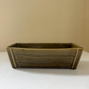 May include: A rectangular olive green ceramic planter with a molded wood grain texture on the exterior. The vessel features vertical corner accents that resemble structural supports, creating a crate-like appearance with a smooth, glossy finish.