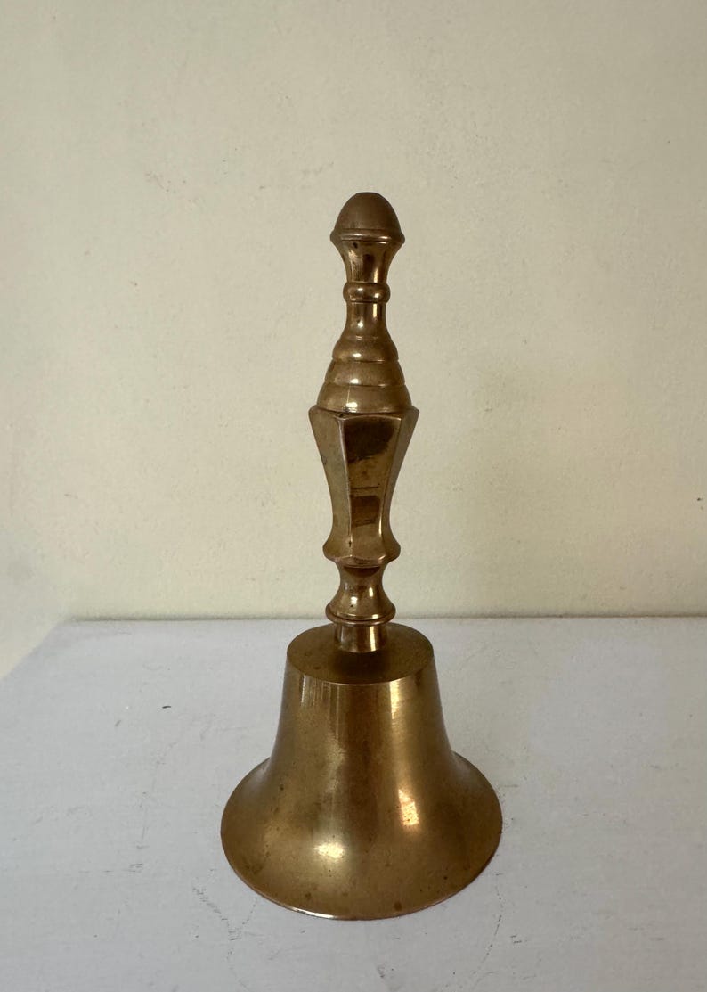 May include: A vintage brass bell with a flared base and a decorative handle. The bell has a warm, golden hue and a classic design, suitable for various decorative purposes. The bell measures approximately 6 inches in height.