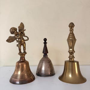 May include: Three antique bells of varying sizes and designs. The bell on the left has a cherub figure on top. The middle bell is silver-toned with a wooden handle. The bell on the right is gold-toned with a long handle.