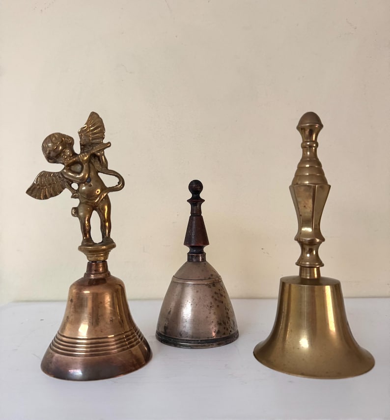 May include: Three antique bells of varying sizes and designs. The bell on the left has a cherub figure on top. The middle bell is silver-toned with a wooden handle. The bell on the right is gold-toned with a long handle.