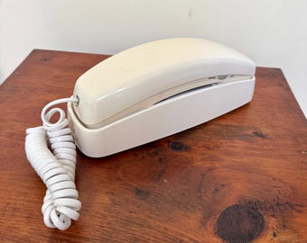 May include: A wall-mountable landline telephone featuring a cream-colored handset resting on a white base. The device includes a coiled white cord extending from the side of the handset, connecting it to the main unit for a functional wired connection.