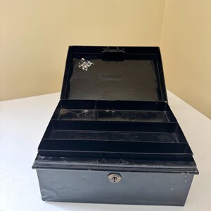 Puede incluir: Caja registradora de metal negro abierta con un ojo de cerradura y compartimentos interiores. La caja est&aacute; abierta, revelando varios compartimentos para organizar efectivo y monedas. La caja est&aacute; sobre una superficie blanca.