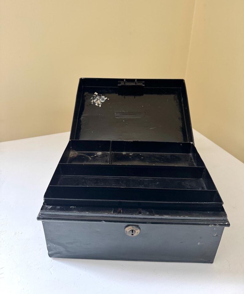 Puede incluir: Caja registradora de metal negro abierta con un ojo de cerradura y compartimentos interiores. La caja est&aacute; abierta, revelando varios compartimentos para organizar efectivo y monedas. La caja est&aacute; sobre una superficie blanca.