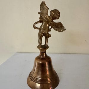 May include: A vintage brass bell featuring a cherub figurine with outstretched wings atop the bell. The bell has a classic shape with horizontal ridges and a warm, golden hue. The cherub is detailed, adding a decorative touch to the piece.