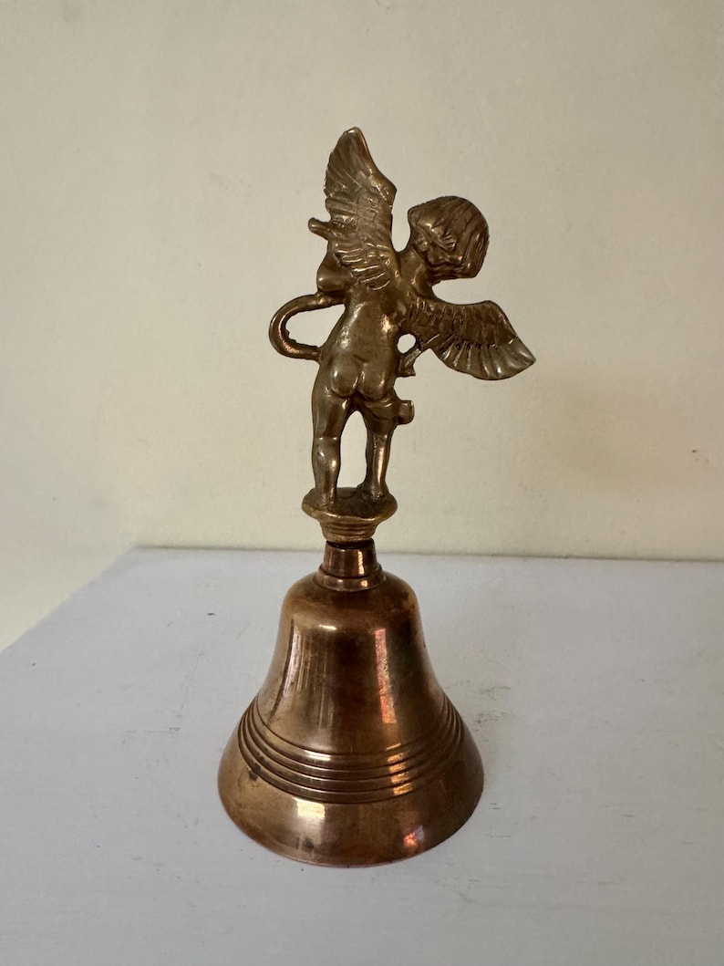 May include: A vintage brass bell featuring a cherub figurine with outstretched wings atop the bell. The bell has a classic shape with horizontal ridges and a warm, golden hue. The cherub is detailed, adding a decorative touch to the piece.