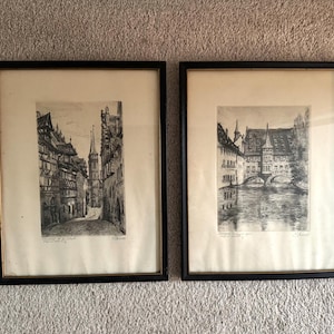 May include: Two framed black and white etchings depicting European cityscapes. The left print shows a narrow street with timber-framed buildings and a church spire. The right print features a stone bridge over a river with historic architecture.