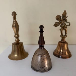 May include: Three antique bells of varying designs. The bell on the left is gold-colored with a long handle. The center bell is silver with a wooden top. The bell on the right features a small angel figure. All three bells are on a white surface.