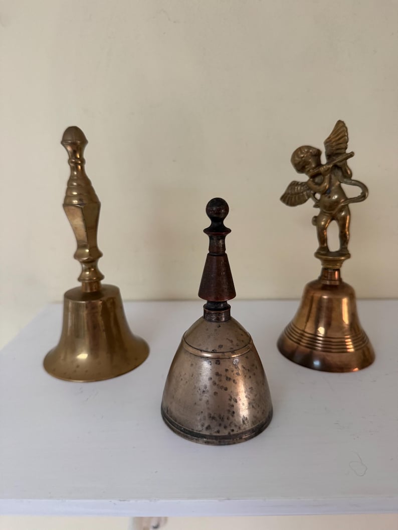 May include: Three antique bells of varying designs. The bell on the left is gold-colored with a long handle. The center bell is silver with a wooden top. The bell on the right features a small angel figure. All three bells are on a white surface.