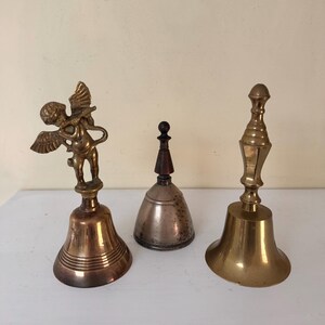 May include: Three antique bells of varying designs. One bell features a cherub figure, another is silver-toned with a wooden handle, and the third is a brass bell with a decorative handle. The bells are displayed on a white surface.
