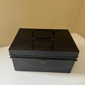 Puede incluir: Una caja registradora de metal negro con forma rectangular. Tiene un asa en la parte superior y un ojo de cerradura en la parte delantera. La caja est&aacute; sobre una superficie blanca.