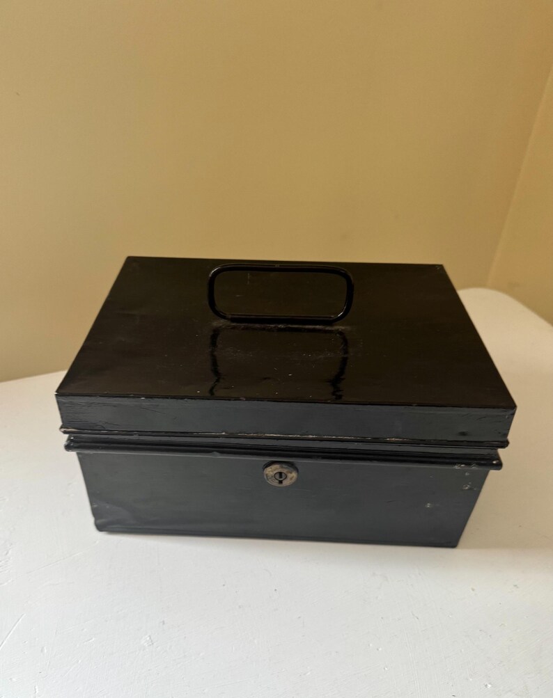 Puede incluir: Una caja registradora de metal negro con forma rectangular. Tiene un asa en la parte superior y un ojo de cerradura en la parte delantera. La caja est&aacute; sobre una superficie blanca.